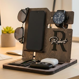 Può includere: Un organizer da scrivania in legno marrone scuro. Dispone di slot per smartphone, occhiali da sole e orologio. L'organizer ha anche un vassoio per piccoli oggetti e il nome "Robert" è visualizzato.