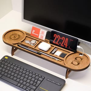 May include: A wooden desk organiser with multiple compartments, holding a phone displaying 22:24, keys, pens, and other small items. The organiser has a circular area with the letter "S" engraved. A keyboard and a cup of coffee are also visible.