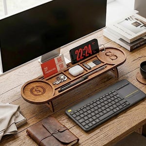 May include: A wooden desk organizer with a monitor stand, featuring compartments for a phone, keys, and accessories. The organizer has a circular coaster with an "S" monogram. A wireless keyboard and a leather-bound notebook are also on the desk.