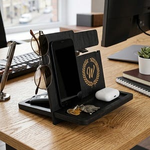 May include: A black wooden desk organizer featuring a vertical stand for a smartphone, a cutout for eyeglasses, and a tray for small items. The front panel displays a gold-colored laurel wreath emblem surrounding the letter W.