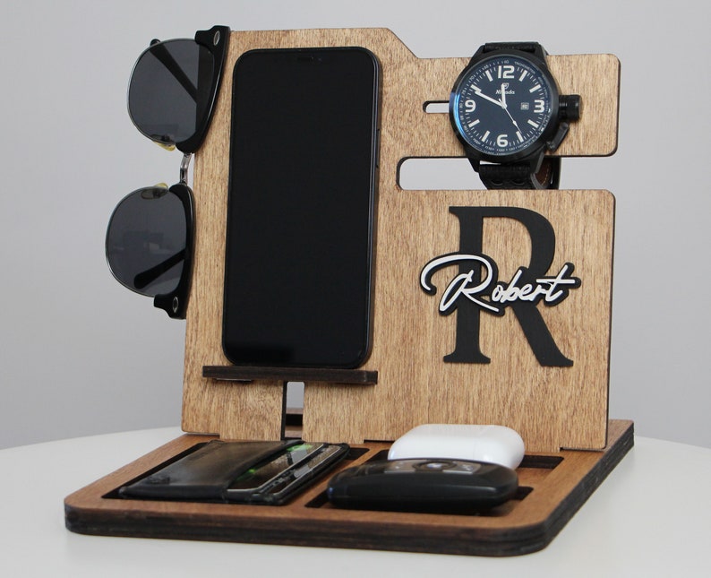 May include: Wooden desk organizer with compartments for a phone, sunglasses, watch, and other accessories. The organizer has a personalized name and initial. The watch has a black band and face. The sunglasses are black.