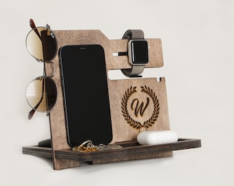 May include: Wooden desk organizer with slots for a smartphone, smartwatch, sunglasses, and small accessories. The organizer is made of dark wood and features a carved initial "W" within a laurel wreath design. Includes a small tray for keys and a pair of white earbuds.