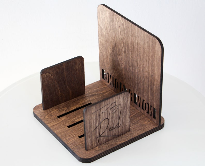 Puede incluir: Un soporte para libros de madera con un tinte marr&oacute;n oscuro. El soporte tiene un dise&ntilde;o recortado que dice "To Be Read" y una ranura para sostener libros.