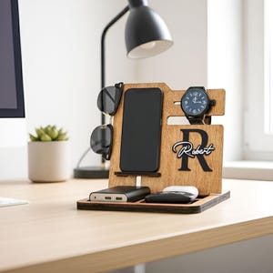 May include: Wooden desk organizer with slots for a phone, sunglasses, and watch. Features a black watch, a black phone, and a power bank. The organizer has the name "Robert" engraved on it. The background includes a desk, a lamp, and a plant.