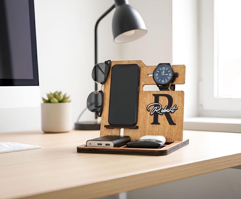 May include: Wooden desk organizer with slots for a phone, sunglasses, and watch. Features a black watch, a black phone, and a power bank. The organizer has the name "Robert" engraved on it. The background includes a desk, a lamp, and a plant.
