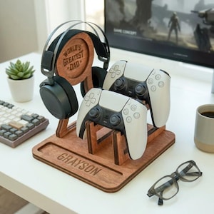 May include: A wooden gaming organiser stand featuring a circular cutout engraved with the text World's Greatest Dad, a base engraved with the name Grayson, a headphone hanger, and two slots for holding white and black wireless game controllers.