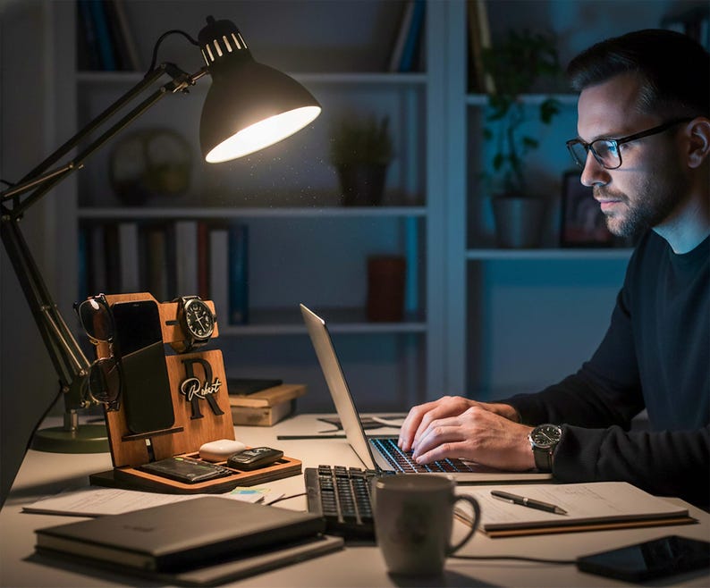 May include: A wooden desk organizer with compartments for a phone, watch, and sunglasses. The organizer has a charging pad and a personalized initial. A black desk lamp illuminates the desk, where a laptop, mug, and notebook are also present.