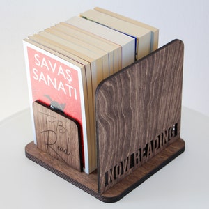 Puede incluir: Un sujetalibros de madera con las palabras "To Be Read" y "Now Reading" grabadas en &eacute;l. El sujetalibros sostiene una pila de libros, uno de los cuales tiene el t&iacute;tulo "Savas Sanati" en la portada.