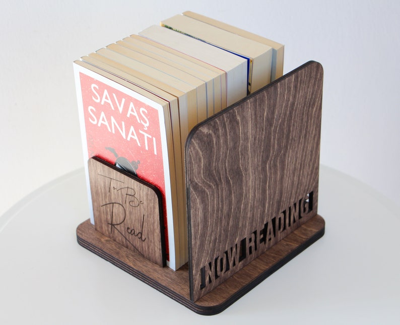 Puede incluir: Un sujetalibros de madera con las palabras "To Be Read" y "Now Reading" grabadas en &eacute;l. El sujetalibros sostiene una pila de libros, uno de los cuales tiene el t&iacute;tulo "Savas Sanati" en la portada.