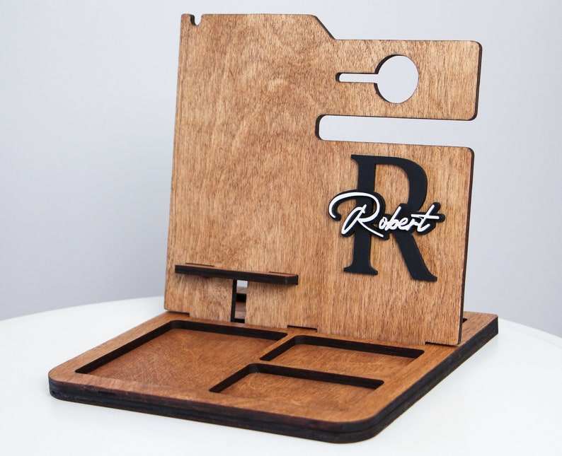 May include: Wooden desk organizer with a phone holder, a small tray, and a cutout for a charging cable. The organizer has a dark brown finish and features the name "Robert" in a cursive font with a large black "R".