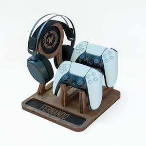 May include: A dark brown wooden gaming stand featuring a circular cutout with a letter W inside a laurel wreath design. The base includes the name FERNANDO engraved in the wood. Two white game controllers and black over-ear headphones are displayed.