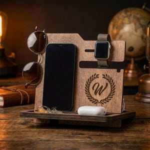 May include: A wooden desk organiser with compartments for a smartphone, sunglasses, a smartwatch, keys, and wireless earbuds. The organiser has a tray at the bottom and a decorative wreath with the letter "W" engraved on the front. The sunglasses are brown.