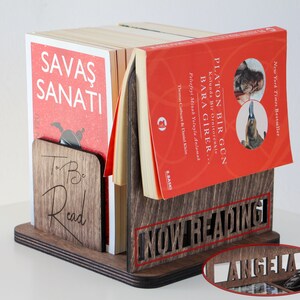 Puede incluir: Un sujetalibros de madera con las palabras "To Be Read" y "Now Reading" grabadas en &eacute;l. El sujetalibros sostiene una pila de libros con cubiertas rojas. El libro de arriba tiene el t&iacute;tulo "Platon Bir G&uuml;n" y el libro de abajo tiene el t&iacute;tulo "Savaş Sanatı".