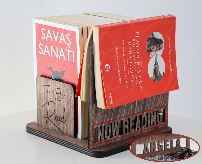 Puede incluir: Un sujetalibros de madera con las palabras "To Be Read" y "Now Reading" grabadas en &eacute;l. El sujetalibros sostiene una pila de libros con cubiertas rojas. El libro de arriba tiene el t&iacute;tulo "Platon Bir G&uuml;n" y el libro de abajo tiene el t&iacute;tulo "Savaş Sanatı".
