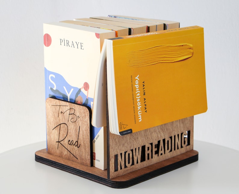 Puede incluir: Un soporte de libros de madera con las palabras "To Be Read" y "NOW READING" grabadas en &eacute;l. El soporte sostiene una pila de libros, incluyendo uno con una cubierta amarilla y el t&iacute;tulo "Yapı(t)s&ouml;k&uuml;m" de YALIN ALPAY.