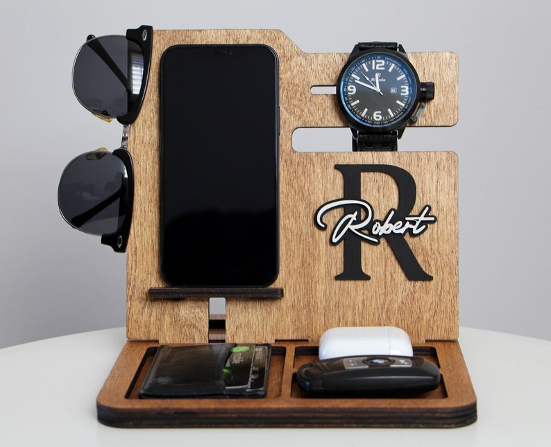 May include: Wooden desk organizer with compartments for a phone, wallet, watch, sunglasses, keys, and wireless earbuds. The organizer has a dark wood finish and features the name "Robert" with a large initial "R".