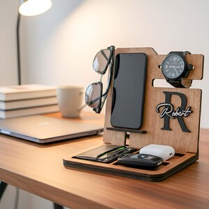 May include: Wooden desk organizer with compartments for a phone, sunglasses, watch, and other accessories. The organizer has a personalized name and initial. A black watch with a black band is displayed on the right side.