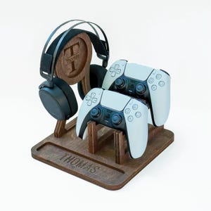 May include: A dark brown wooden gaming stand featuring a circular cutout with the letter T and the name Thomas engraved. The stand holds a pair of black over-ear headphones and two white wireless game controllers, with the name Thomas engraved on the base.