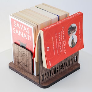 Puede incluir: Un sujetalibros de madera con las palabras "NOW READING" talladas en la parte delantera. El sujetalibros sostiene una pila de libros, incluyendo uno con el t&iacute;tulo "PLATON BIR GUN BARAGIRER" en turco. El sujetalibros tambi&eacute;n tiene las palabras "To Be Read" talladas en el lateral.