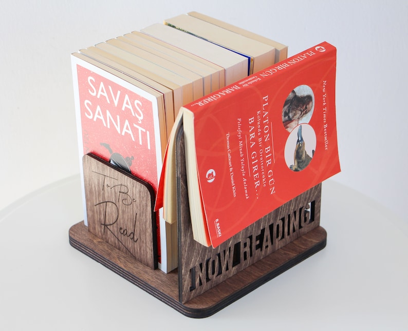 Puede incluir: Un sujetalibros de madera con las palabras "NOW READING" talladas en la parte delantera. El sujetalibros sostiene una pila de libros, incluyendo uno con el t&iacute;tulo "PLATON BIR GUN BARAGIRER" en turco. El sujetalibros tambi&eacute;n tiene las palabras "To Be Read" talladas en el lateral.