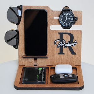 May include: Wooden desk organizer with compartments for a phone, wallet, watch, sunglasses, keys, and earbuds. The organizer is engraved with the name "Robert" and a large letter "R". The watch has a black face and a black leather band.