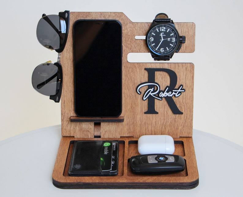 May include: Wooden desk organizer with compartments for a phone, wallet, watch, sunglasses, keys, and earbuds. The organizer is engraved with the name "Robert" and a large letter "R". The watch has a black face and a black leather band.