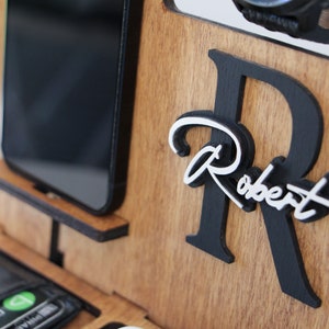 May include: Wooden desk organizer with compartments for a phone, wallet, watch, and glasses. Features a black and white personalized name, "Robert", on the front. The organizer is made of light brown wood.