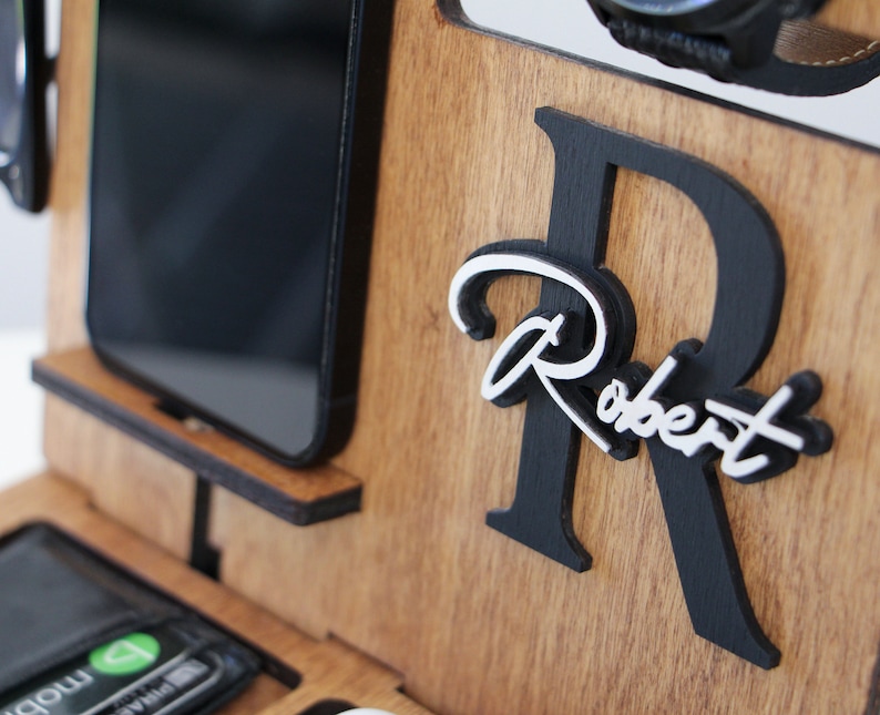 May include: Wooden desk organizer with compartments for a phone, wallet, watch, and glasses. Features a black and white personalized name, "Robert", on the front. The organizer is made of light brown wood.