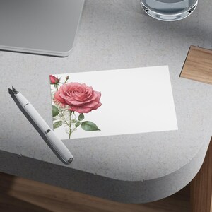 May include: A white card featuring a watercolor illustration of a pink rose with green leaves and white accents. A silver and white pen is positioned near the card. The card is on a gray surface.