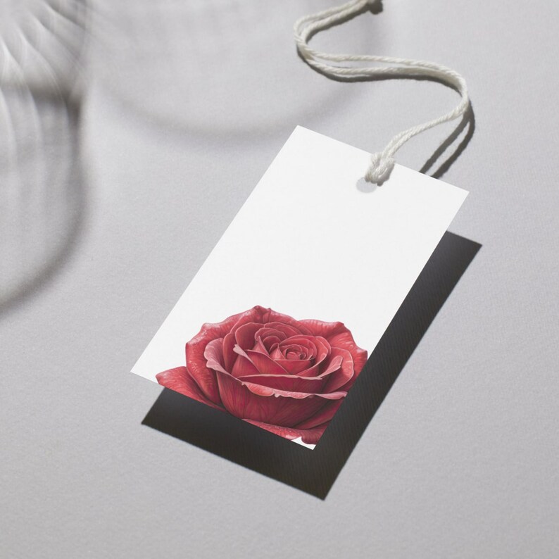 May include: A white rectangular tag with a red rose illustration at the bottom. The tag is attached to a white string. The rose is a deep red color with detailed petals, and the tag casts a shadow on the gray surface.