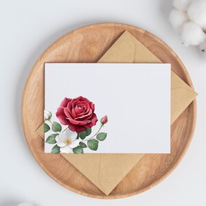 May include: A white card with a watercolor design of a red rose and white flowers, with green leaves and buds. The card is on a wooden tray with a brown envelope.
