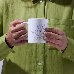 May include: White ceramic mug featuring a lavender floral design in shades of purple and green. The mug has a curved handle and is held by a person wearing a green shirt. The text at the bottom of the image reads: EXAMPLE OF HOW THEY CAN BE USED.