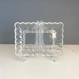 May include: A clear glass serving dish with a scalloped edge and a raised inner border. The dish is on a clear plastic stand.