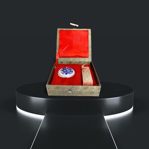 May include: A green and gold box with a red interior, containing a white and blue porcelain inkwell and a carved wooden stamp. The box is on a black pedestal.