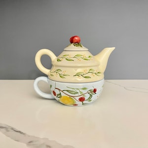 May include: A white ceramic teapot with a blue and green floral design. The teapot has a lid with a red cherry on top. The teapot is sitting on a white surface.