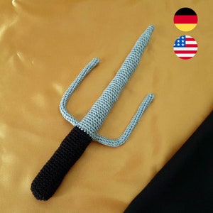 May include: A crocheted gray and black sai, a traditional Japanese weapon, with three prongs. The sai is lying on a gold fabric surface.