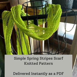 May include: A vibrant green, knitted scarf with a striped design is displayed on a black metal stand. The scarf features an open, airy weave. The image includes the text "Simple Spring Stripes Scarf Knitted Pattern".