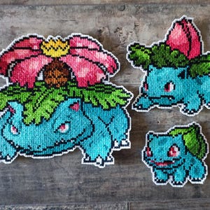 Op de afbeelding: Drie pixel art Bulbasaur figuren in verschillende maten. De grootste heeft een roze en gele bloem op de rug, de middelste heeft een roze knop en de kleinste heeft een groen blad. Alle zijn turquoise met rode ogen.