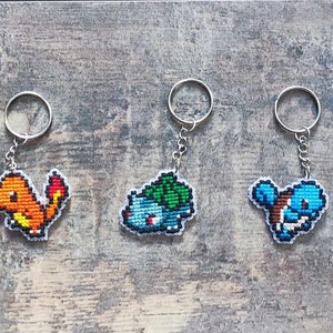 Könnte beinhalten: Drei Schlüsselanhänger mit gestickten Pixel-Pokémon-Charakteren. Der Schlüsselanhänger links zeigt einen roten und orangefarbenen Glurak, der mittlere Schlüsselanhänger zeigt einen grünen und blauen Bisasam, und der Schlüsselanhänger rechts zeigt einen blauen Schiggy.