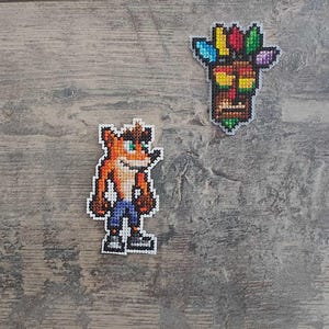 Op de afbeelding: Borduurwerk met kruissteek van een cartoonfiguur, Crash Bandicoot, in oranje, bruin, blauw en wit. Een tweede borduurwerk met kruissteek van een tiki-masker met een kleurrijke hoofdtooi is ook te zien.