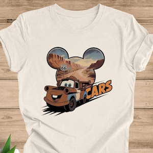 May include: Off-white t-shirt featuring a cartoon image of Mater from the movie Cars. The design includes a Route 66 sign and the word "Cars" in orange. The background is a desert landscape.