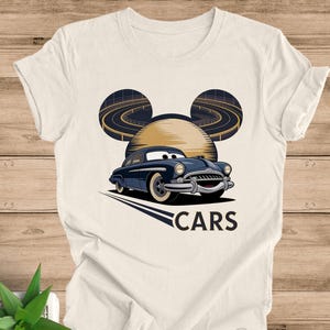 May include: Cream-colored t-shirt featuring a cartoon car design. The design includes a navy blue car with white tires, set against a backdrop of a stylized sun and race tracks, with the word "CARS" below.