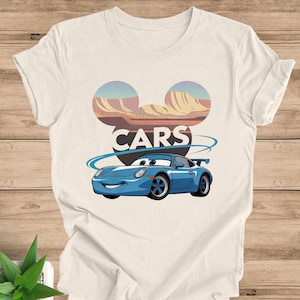 May include: Off-white t-shirt featuring a cartoon car design. The design includes a blue car with a smiling face, the word "CARS" in bold letters, and a stylized mountain range in the background. The shirt is made of a soft material.