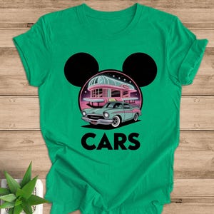 May include: Green t-shirt featuring a retro car design with a diner and the word "CARS" in bold letters. The design includes a light blue car, a pink diner, and black Mickey Mouse ears.