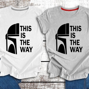 May include: Two t-shirts featuring a black and white graphic of a Mandalorian helmet and the text "This is the way". One t-shirt is white and the other is grey.