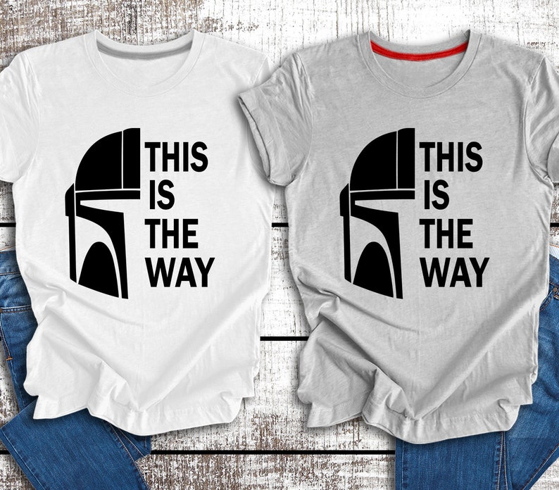 May include: Two t-shirts featuring a black and white graphic of a Mandalorian helmet and the text "This is the way". One t-shirt is white and the other is grey.