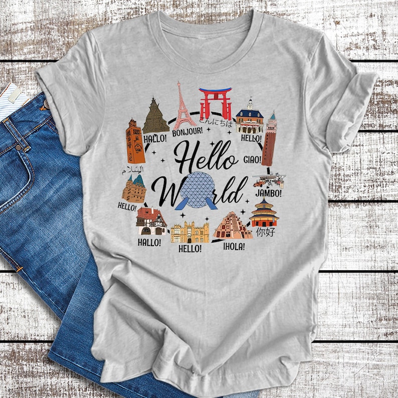 May include: A gray t-shirt with a graphic of the Spaceship Earth ride at Epcot, surrounded by iconic landmarks from around the world. The text "Hello World" is written in a circle around the Spaceship Earth, with greetings in different languages.