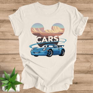 May include: Off-white t-shirt featuring a cartoon car design. The design includes a blue car with a smiling face, the word "CARS" in bold letters, and a stylized mountain range in the background. The shirt is made of a soft material.