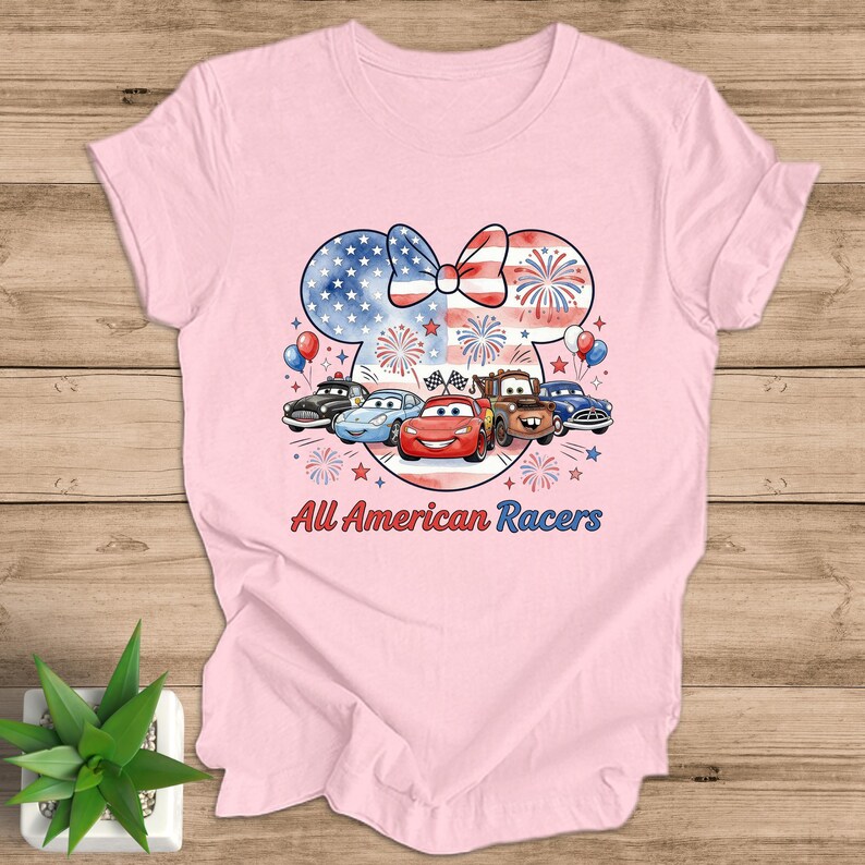 May include: A light pink t-shirt featuring a graphic of a Minnie Mouse head with a bow in the colors of the American flag. Below the head are cartoon cars and the text "All American Racers".