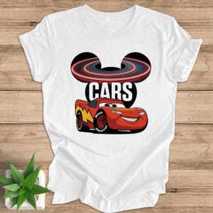 May include: White t-shirt featuring a graphic of Lightning McQueen from the movie Cars. The design includes the word "CARS" and a stylized racetrack. The car is red with yellow and black accents.